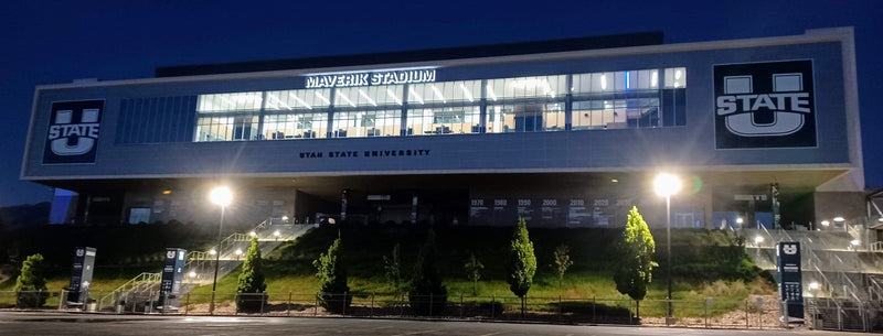 The West Stadium Center at Maverik Stadium in Logan, lit up at dusk, seen from the parking lot
