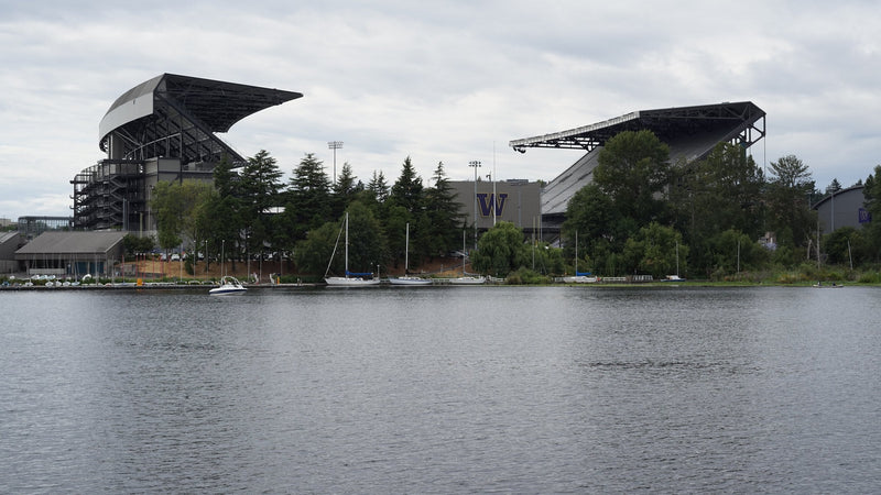 Husky Stadium's two grandstands seen across Union Bay, with sailboats moored at the shore