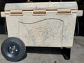 Load image into Gallery viewer, Tan speckled hard cooler on two black knobby wheels and a black stand, seen from the front in a shop