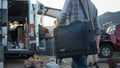 Load image into Gallery viewer, Man carrying a black kitchen bag by its shoulder strap past an open camper van at dusk