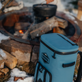 Load image into Gallery viewer, Slate-blue backpack cooler beside a campfire ring with a Dutch oven over burning logs