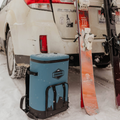 Load image into Gallery viewer, Slate-blue backpack cooler in the snow behind an SUV, with skis leaning on the bumper
