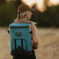 Load image into Gallery viewer, Woman in a ball cap wearing a slate-blue backpack cooler in a golden field at sunset