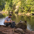 Load image into Gallery viewer, Man crouched by a lake opening a black backpack cooler on the rocky shore, a dog nearby