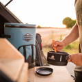 Load image into Gallery viewer, Slate-blue backpack cooler on a tailgate table while someone stirs a camp pot beside it at sunset
