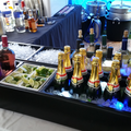 Load image into Gallery viewer, Black cooler at an outdoor event bar holding sparkling wine and wine bottles on ice beside a tray of cut limes