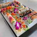 Load image into Gallery viewer, Close angle of sushi rolls, nigiri, pickled ginger and purple orchids on white boards over a black cooler