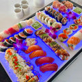 Load image into Gallery viewer, Close-up of sushi and sashimi on white boards glowing blue-purple from cubes in the ice below