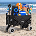 Load image into Gallery viewer, Black folding wagon with gray balloon wheels on beach sand, loaded with a speaker, cooler, beach chair, towels and umbrella