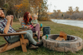 Load image into Gallery viewer, Family in wooden chairs around a stone fire pit by a river, a dark backpack cooler next to them