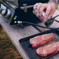 Load image into Gallery viewer, Hand seasoning two raw steaks on a tray beside the grill's control panel and gas hose