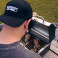 Load image into Gallery viewer, Man in a ball cap cooking two steaks on a tabletop grill set on a picnic table outdoors