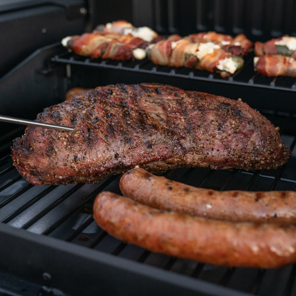 Grilled tri-tip with a temperature probe in it on a tailgate grill, sausages beside it and jalapeño poppers on the upper rack