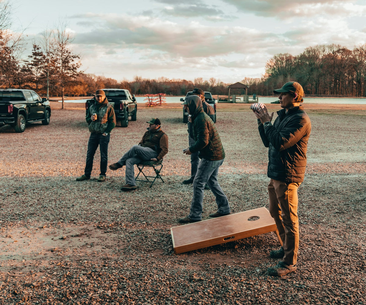 Friends playing cornhole on a gravel lot between parked pickup trucks