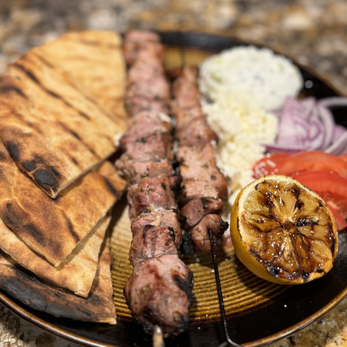 Two pork souvlaki skewers with grilled pita, tzatziki, feta, red onion, tomato and a grilled lemon on a plate