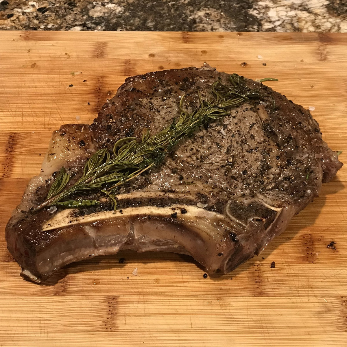 A thick bone-in ribeye with a dark crust, cracked pepper, flaky salt and a rosemary sprig on a cutting board