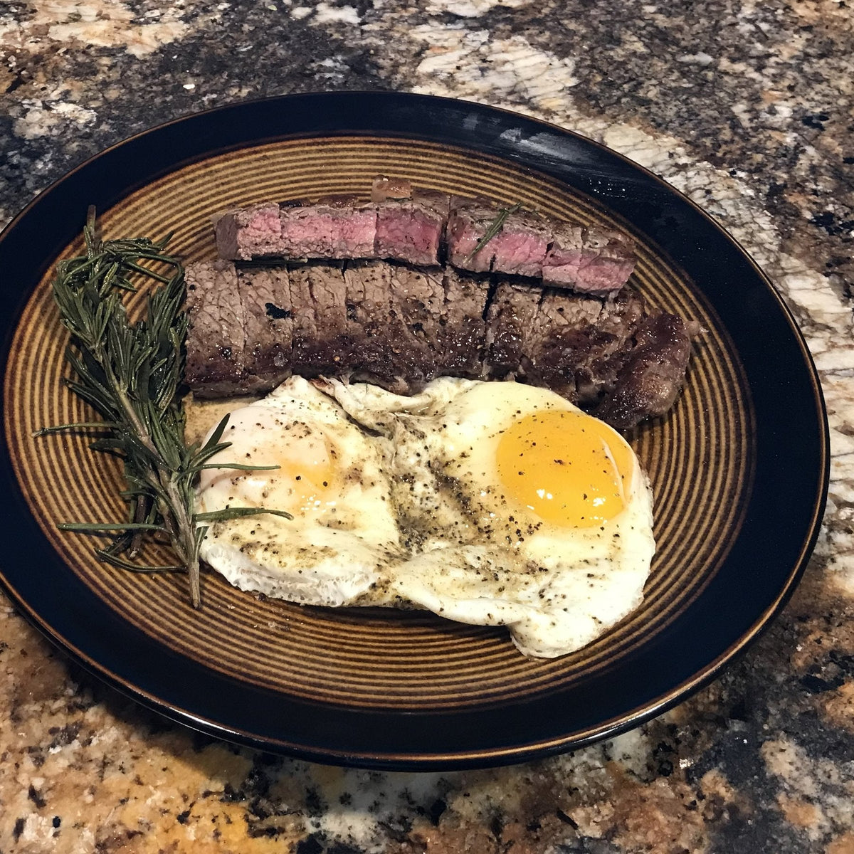A sliced NY strip steak with two sunny-side-up eggs and a rosemary sprig on a plate