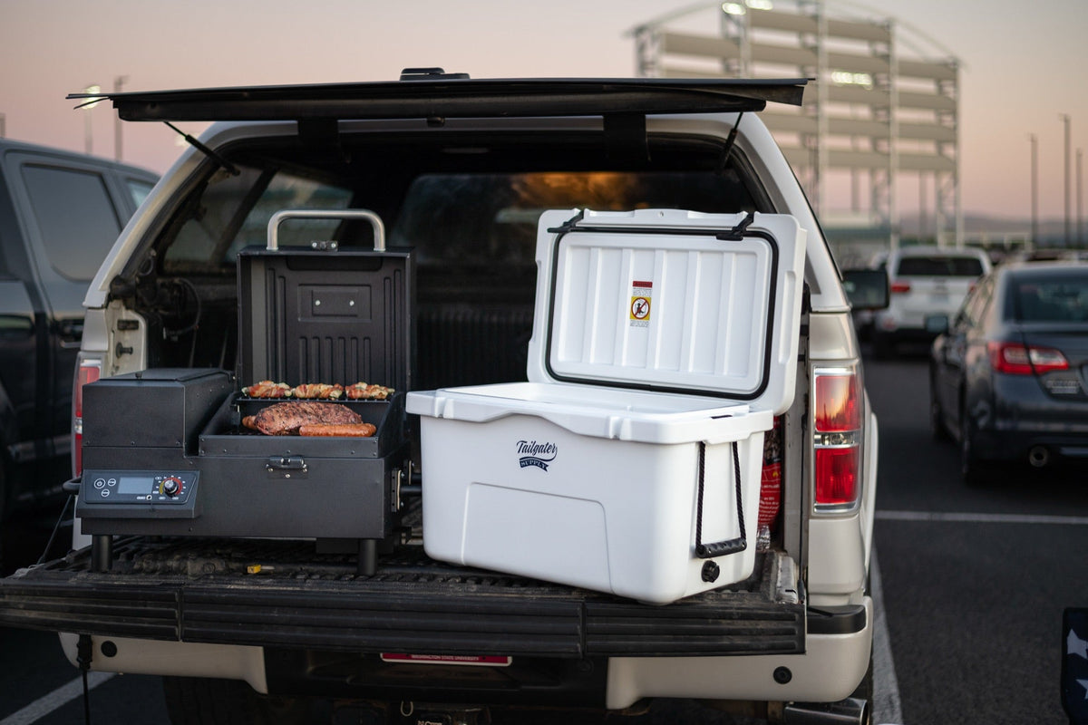 A grill and a white cooler set up in a pickup bed outside a stadium at dusk