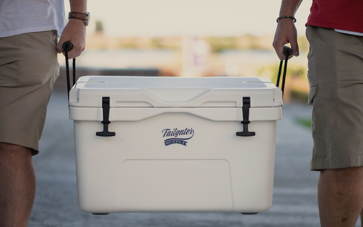 Two people carrying a white hard cooler by its rope handles across a lot