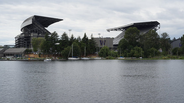 Husky Stadium's two grandstands seen across Union Bay, with sailboats moored at the shore