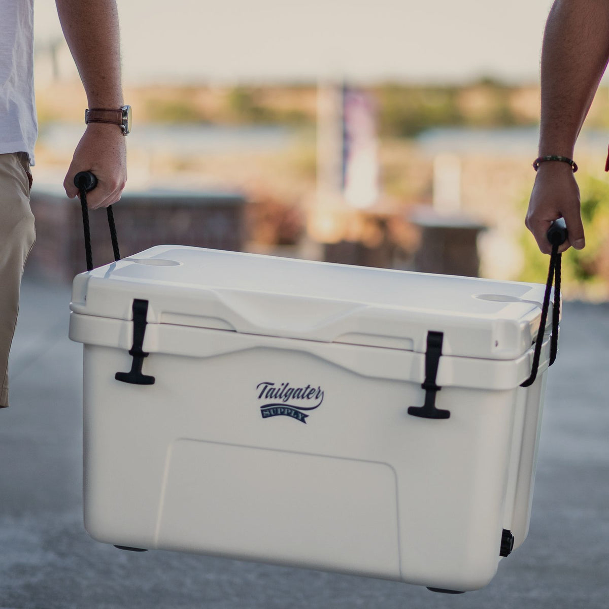 Two people carrying a white Tailgater Supply hard cooler across a lot