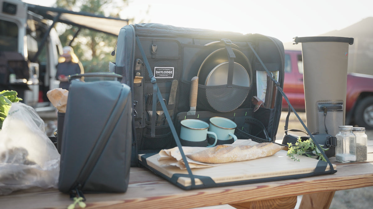 Open camp kitchen bag on a picnic table with the bamboo board folded out, mugs, plates and bread, a camper van behind