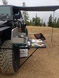 Load image into Gallery viewer, Tire Table on a Jeep's tire under an awning, set up with a camp stove, a cutting board and food prep.
