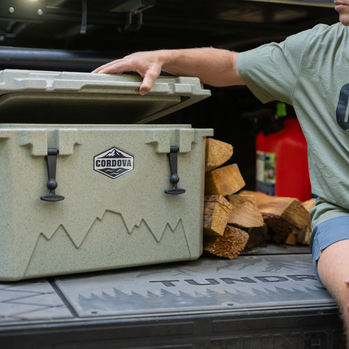 Man opening a tan hard cooler on a pickup tailgate, with split firewood stacked beside it