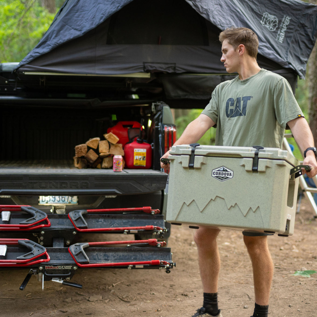 Man carrying a tan hard cooler by its side handles away from a truck with a rooftop tent