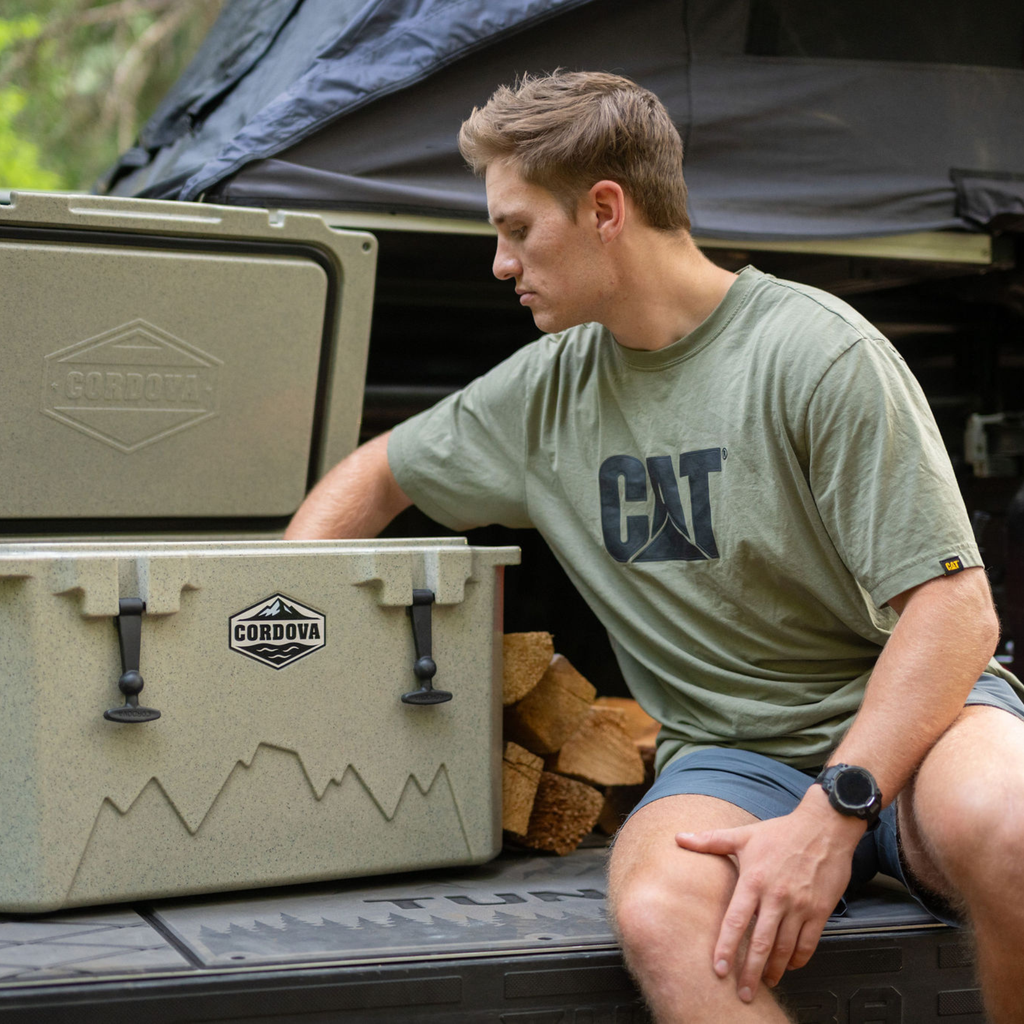 Man sitting on a pickup tailgate next to a tan hard cooler and a stack of firewood