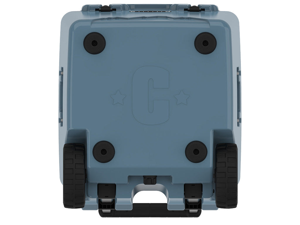 Underside of the slate-blue wheeled cooler showing four rubber feet, two wheels and a molded C logo