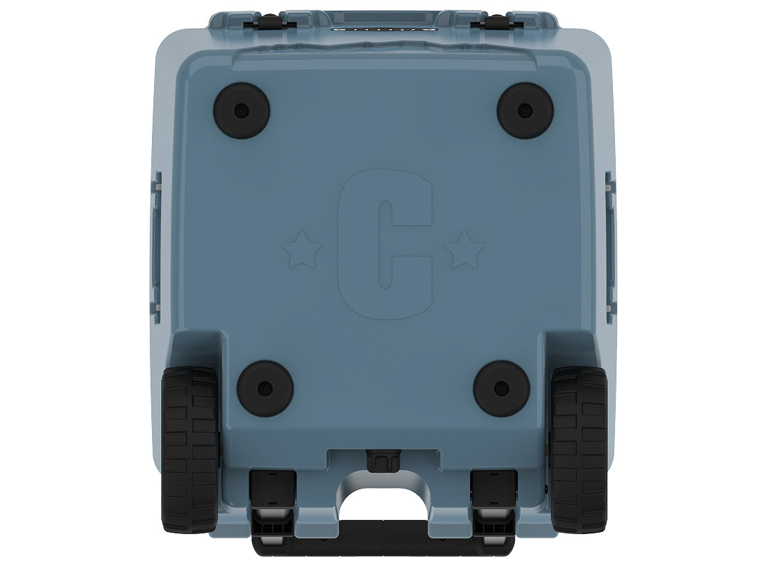 Underside of the slate-blue wheeled cooler showing four rubber feet, two wheels and a molded C logo