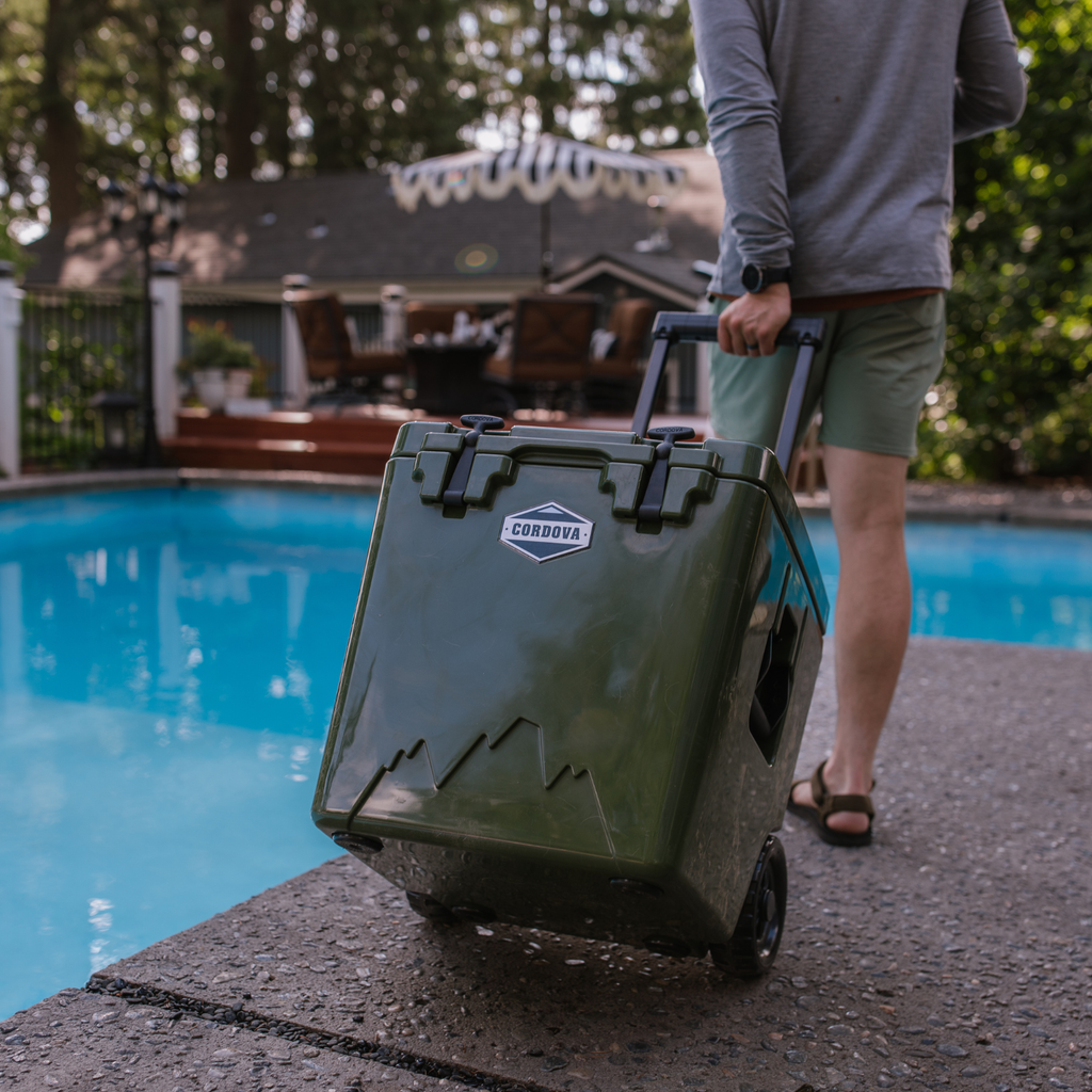 Man rolling an olive green wheeled cooler by its handle along a pool deck