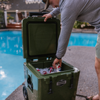 Man lifting the lid of an olive green wheeled cooler packed with soda cans on ice by a pool