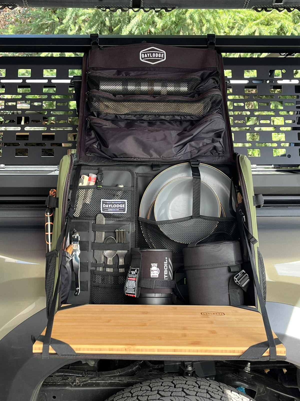 Kitchen bag hung from a truck bed rack, open, with plates, utensils and a stove inside and the board folded out
