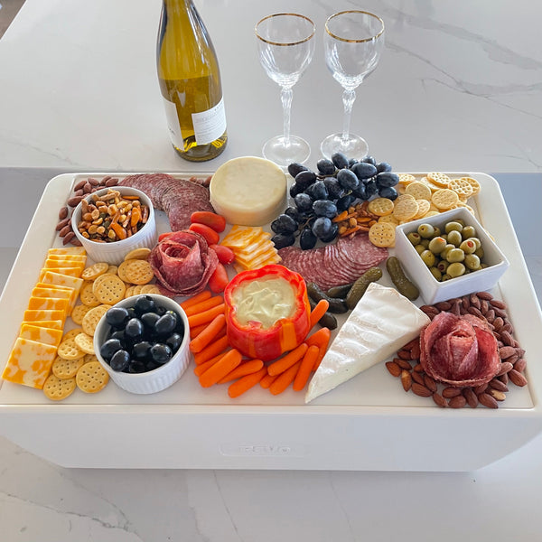 Full charcuterie board covering a white cooler on a marble island, with white wine and two gold-rim glasses