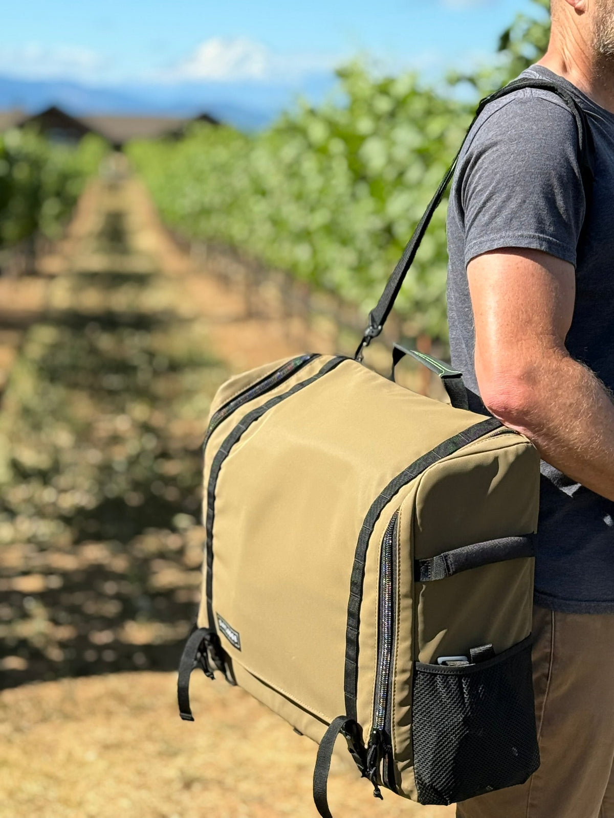 Man carrying a brown camp kitchen bag on a shoulder strap down a vineyard row