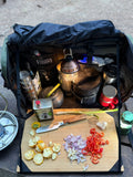 Load image into Gallery viewer, Open kitchen bag on a camp table with chopped peppers and onions on the bamboo board, a lantern and spices inside
