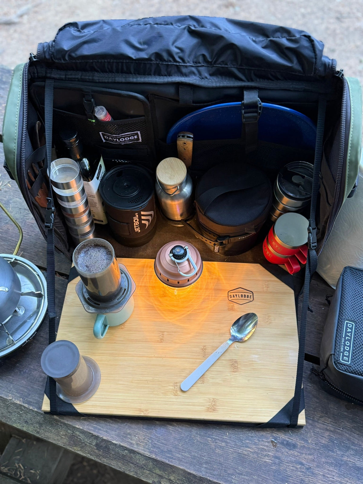 Kitchen bag set up as a coffee station on a picnic table, with a pour-over brewer and a lantern on the board