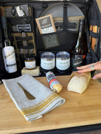 Bamboo board laid with bread, cheese, a towel and a butter knife in front of the open bag's bottles and cups