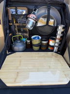 Open kitchen bag packed with a kettle, jars, spice tins, a lantern and plates above an empty bamboo board