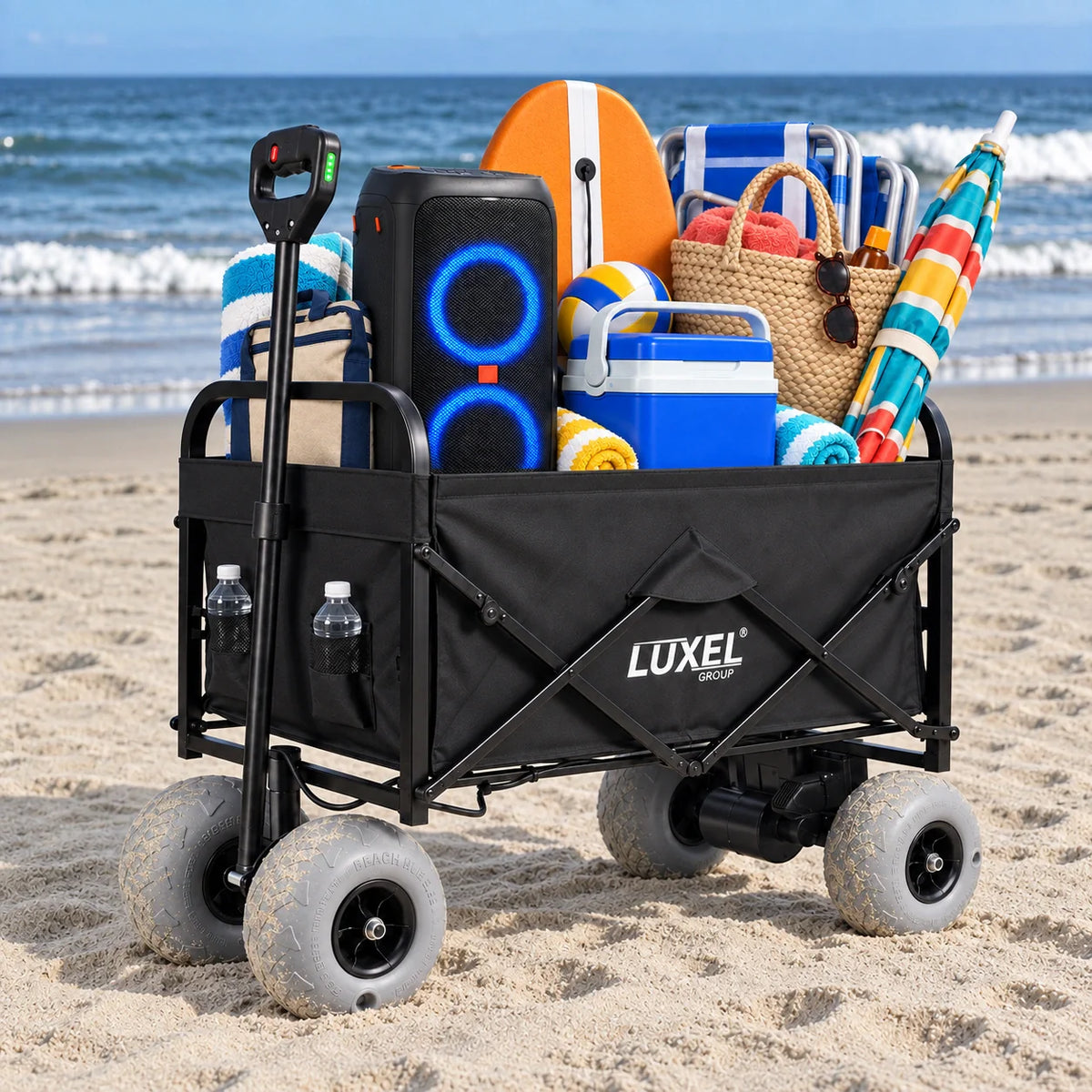 Black folding wagon with gray balloon wheels on beach sand, loaded with a speaker, cooler, beach chair, towels and umbrella