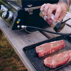 Hand seasoning two raw steaks on a tray beside the grill's control panel and gas hose
