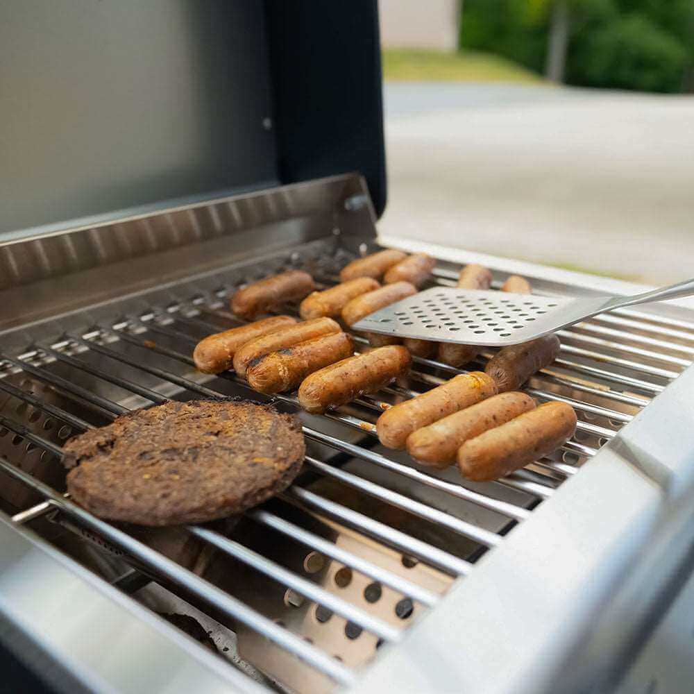 Spatula over sausages and a burger patty cooking on stainless grates, lid open