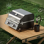 Tabletop grill on a slatted camp table in the grass, hooked to a 1 lb propane cylinder by a short hose