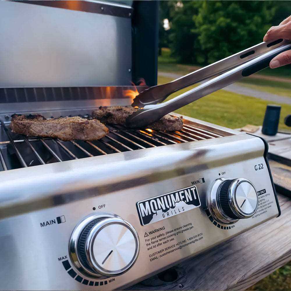 Tongs lifting steaks off the grates, seen past the two control knobs and Monument logo