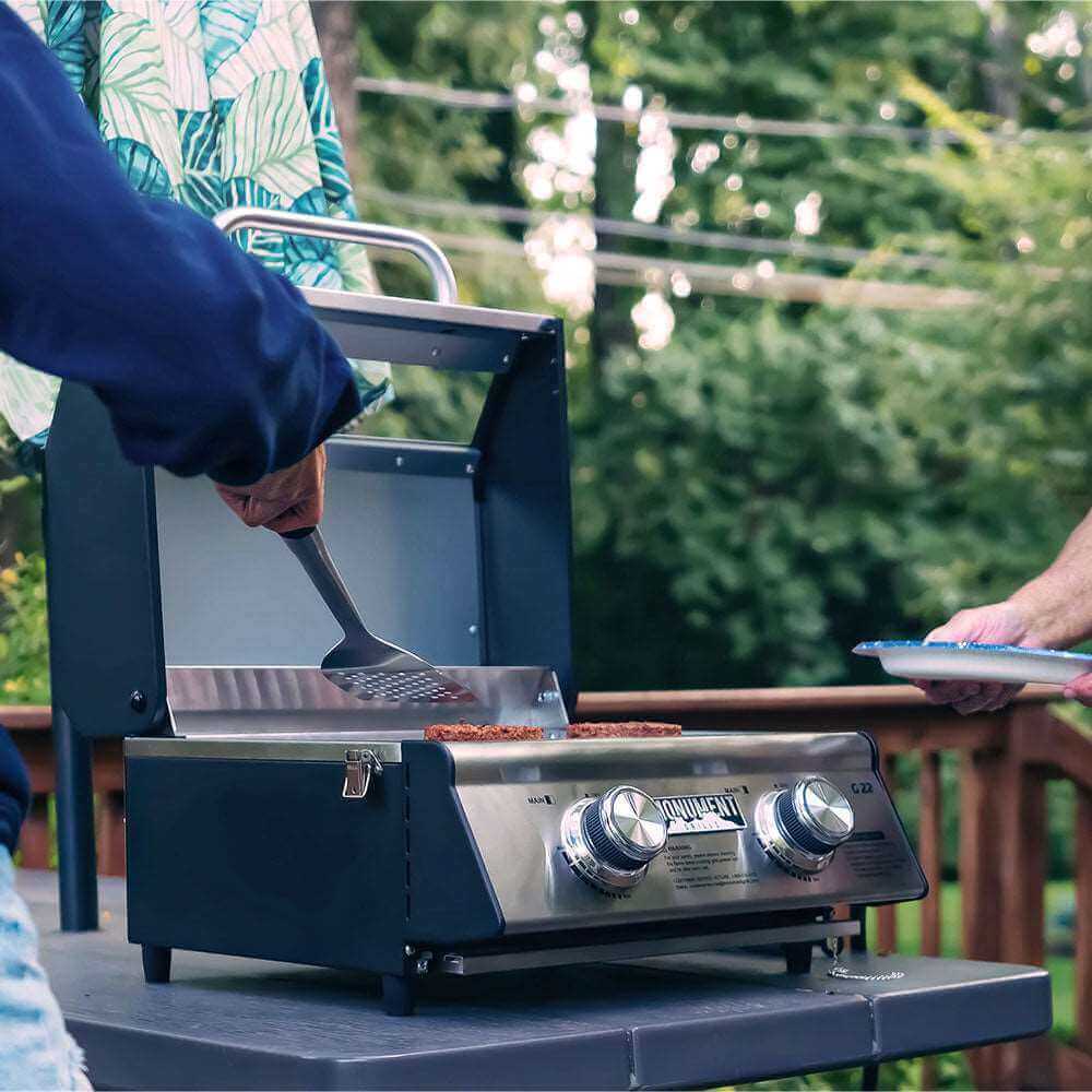 Two people at a deck table, one sliding a spatula under burgers on the open grill