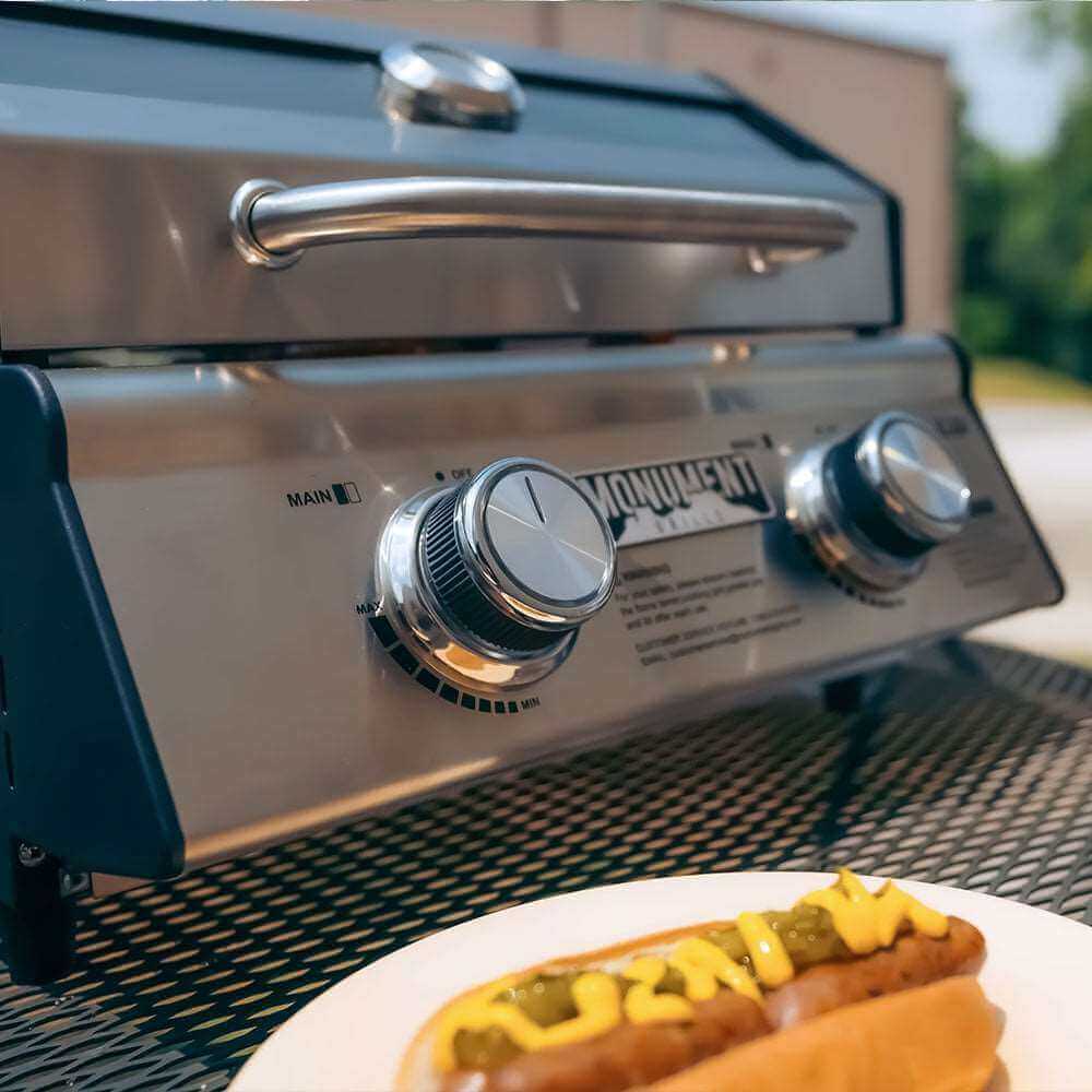 Closed tabletop grill on a mesh patio table with a loaded hot dog on a plate in front