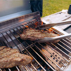 Tongs turning a steak with grill marks on stainless grates over open flame