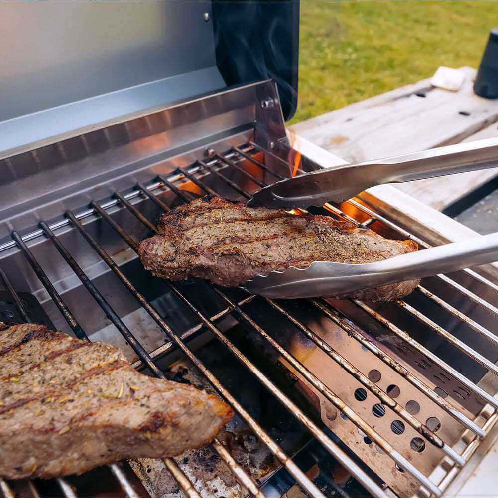 Tongs turning a steak with grill marks on stainless grates over open flame
